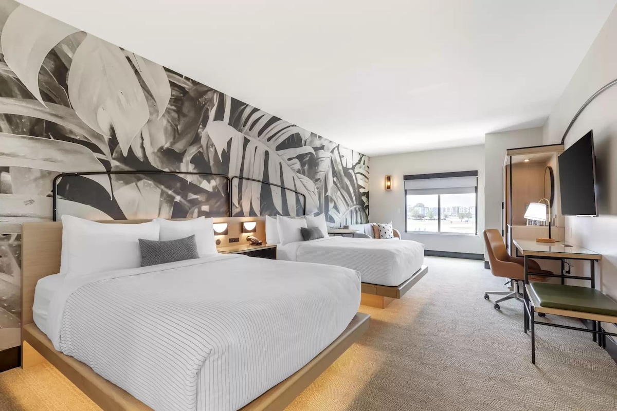 A hotel room with two beds, a desk, a chair, a large wall mural, and a TV.