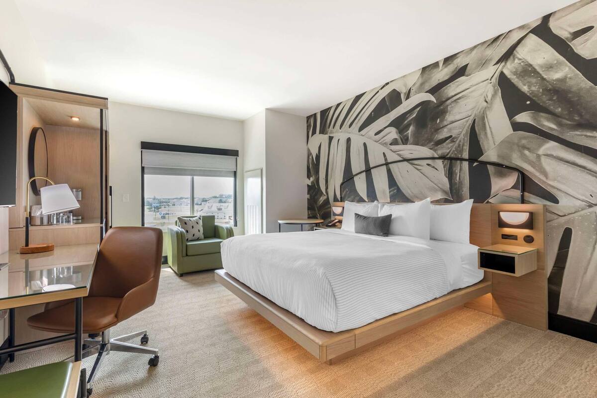 A modern hotel room with a large bed, a desk with a chair, a green sofa, and a large abstract wall painting.