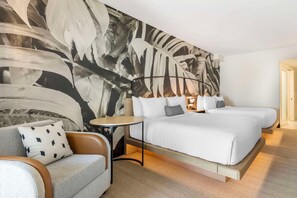A modern hotel room with a large abstract mural on the wall, a sofa with a patterned pillow, a small round table, and two beds with white linens.