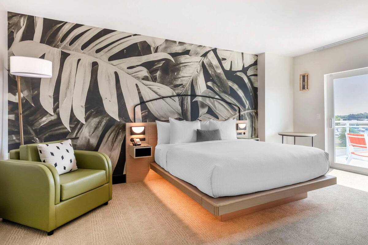 A modern hotel room with a large bed, a green armchair, a nightstand with a lamp, and a large abstract wall mural.
