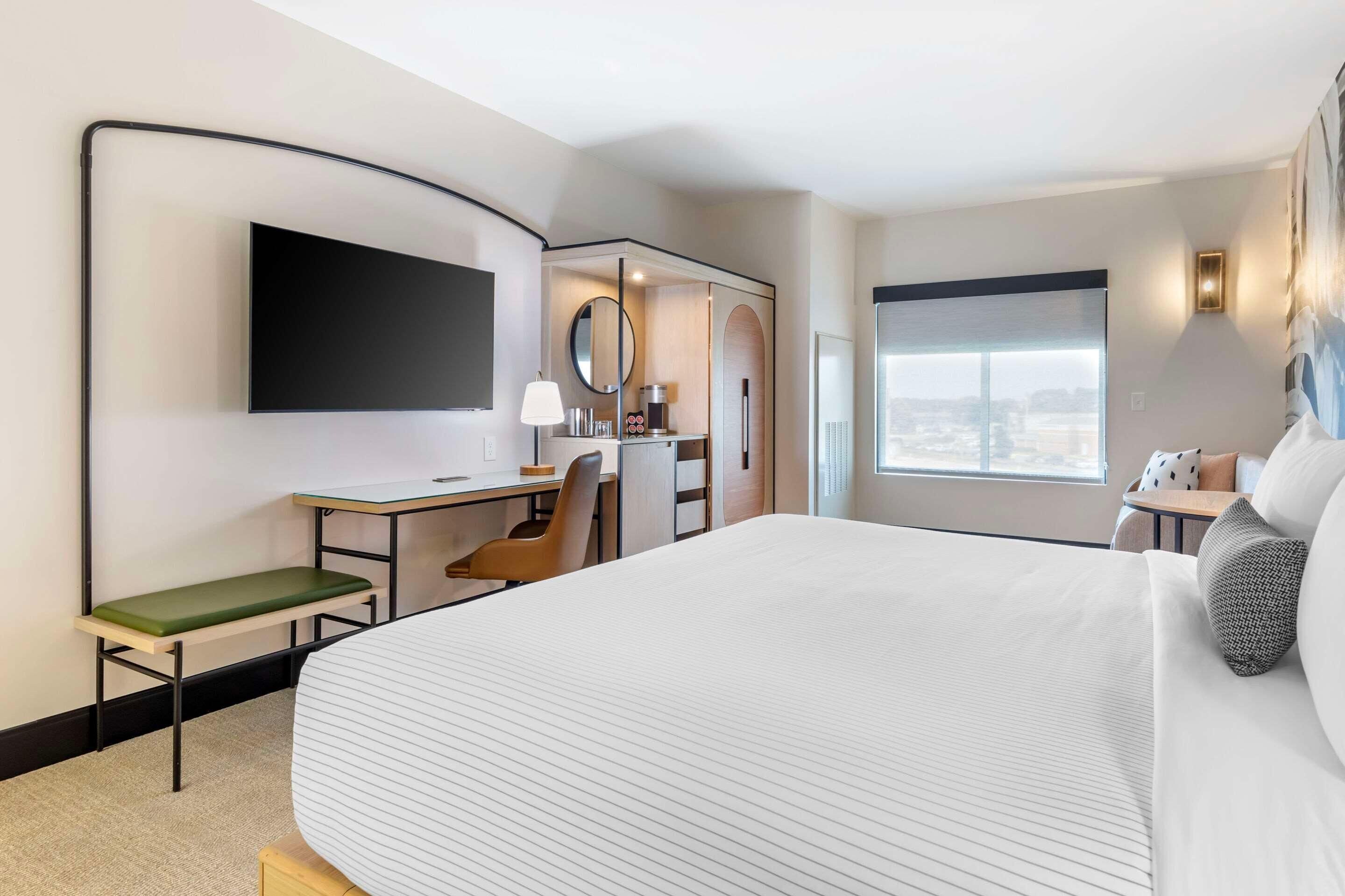 Ein Hotelzimmer mit einem großen Bett, einem Schreibtisch mit Stuhl, einem Fernseher, einem Fenster mit Aussicht und einem kleinen Tisch mit Lampe.