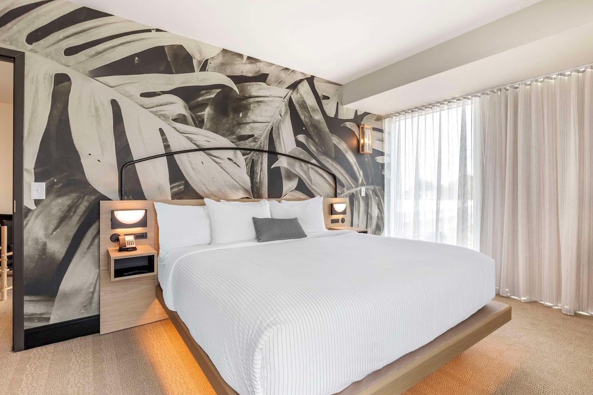 A modern hotel room with a large bed, two bedside tables with lamps, a wall mural, and a large window with curtains.