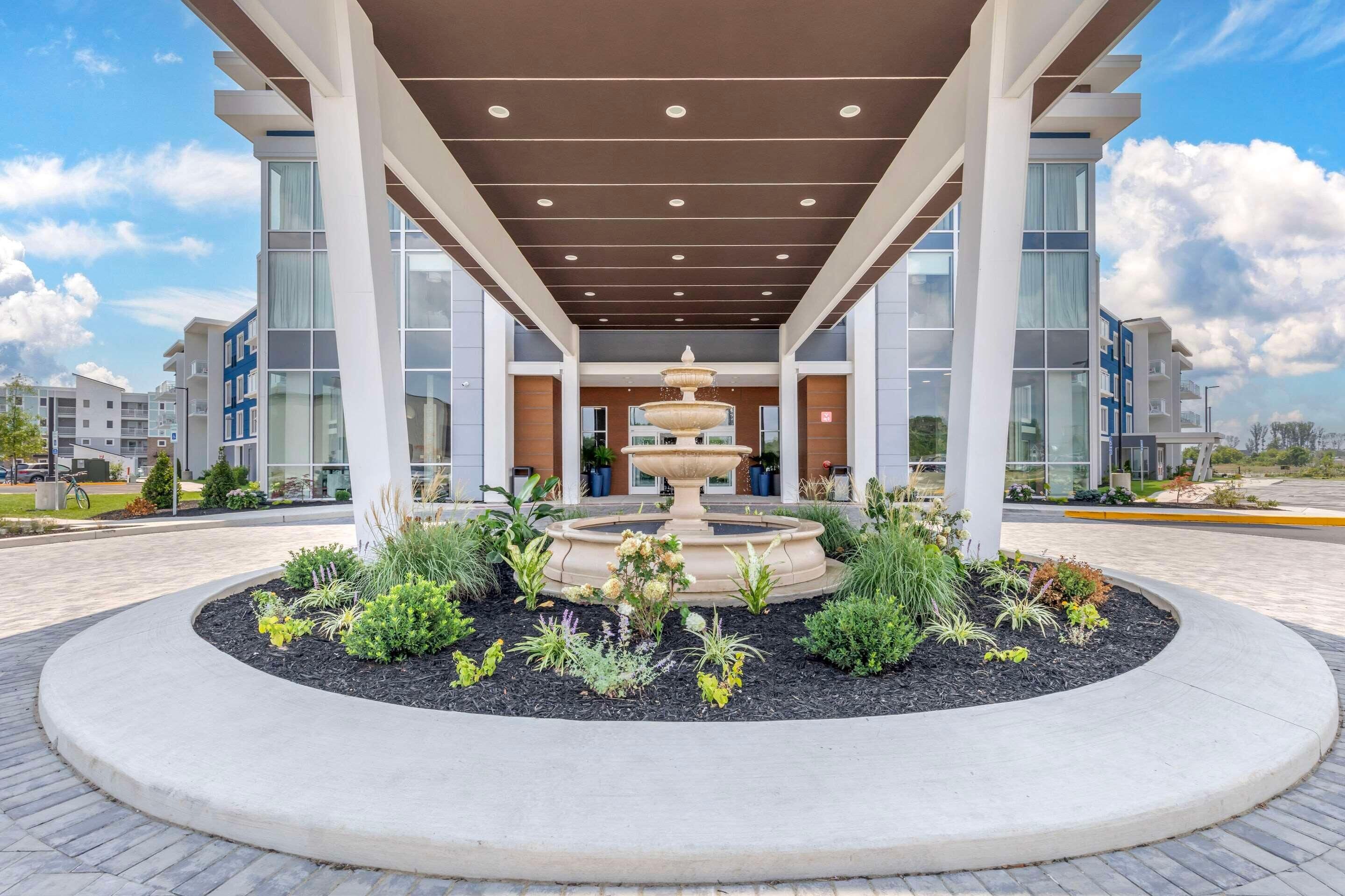 A modern building with a central fountain, surrounded by landscaped garden beds, under a covered walkway.