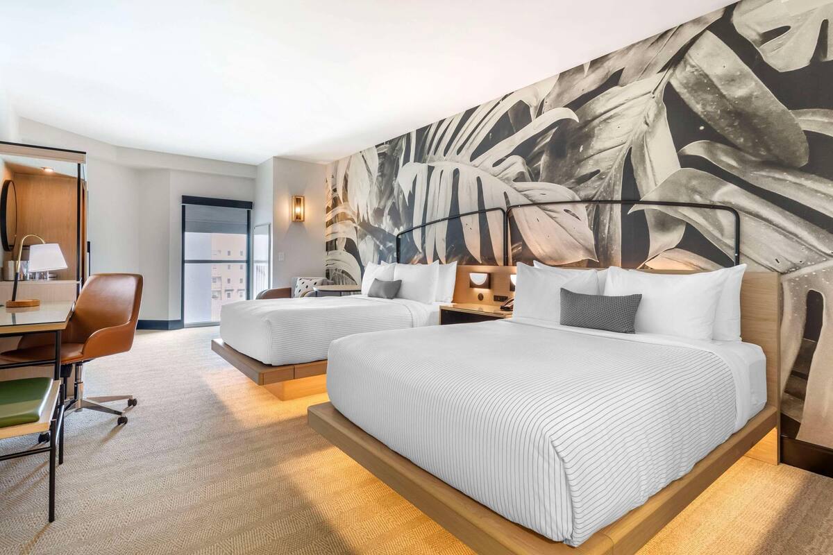 A hotel room with two beds, a desk, a chair, and a large leaf-patterned wall.