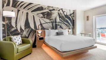 Habitación de hotel moderna con una cama grande, un sillón verde, una mesita de noche con una lámpara y un amplio mural abstracto en la pared.
