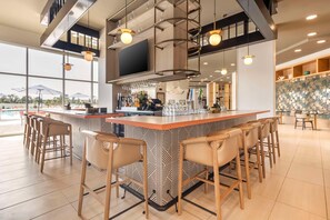 A modern bar area with a patterned bar counter, multiple bar stools, and a large window offering a view of the outdoors. - Cambria Hotel Rehoboth Beach (Rehoboth Beach)