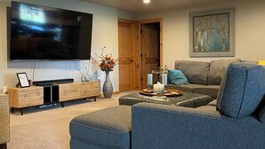 A modern living room with a large flat-screen TV, a grey sectional sofa, a wooden entertainment unit, and a framed artwork on the wall. - Entertainer's Dream - Quick drive to Lambeau - Hot Tub, Bar, & 85" TV. (Green Bay)