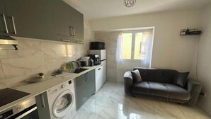 A compact kitchen with a washing machine, a microwave, and a coffee maker. There is a sofa in the living area and a window providing natural light. - Appartement Calme Moderne Proche de Disneyland Wifi & Café Gratuit (Bussy-Saint-Georges)