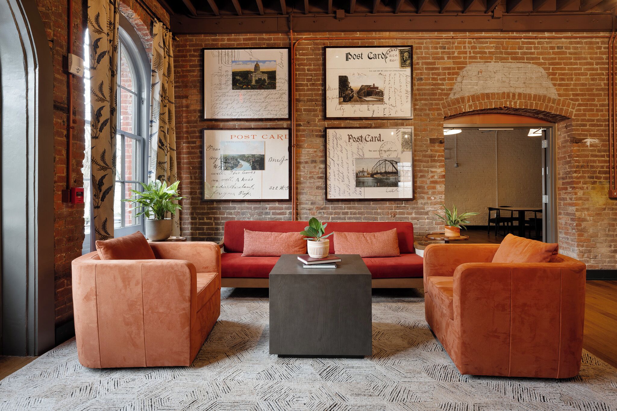 Una habitación acogedora con pared de ladrillo, un sofá rojo, dos sillones naranjas, una mesa de centro y postales enmarcadas en la pared.