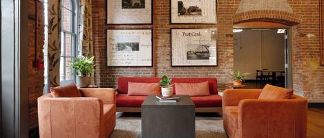 Una habitación acogedora con pared de ladrillo, un sofá rojo, dos sillones naranjas, una mesa de centro y postales enmarcadas en la pared.