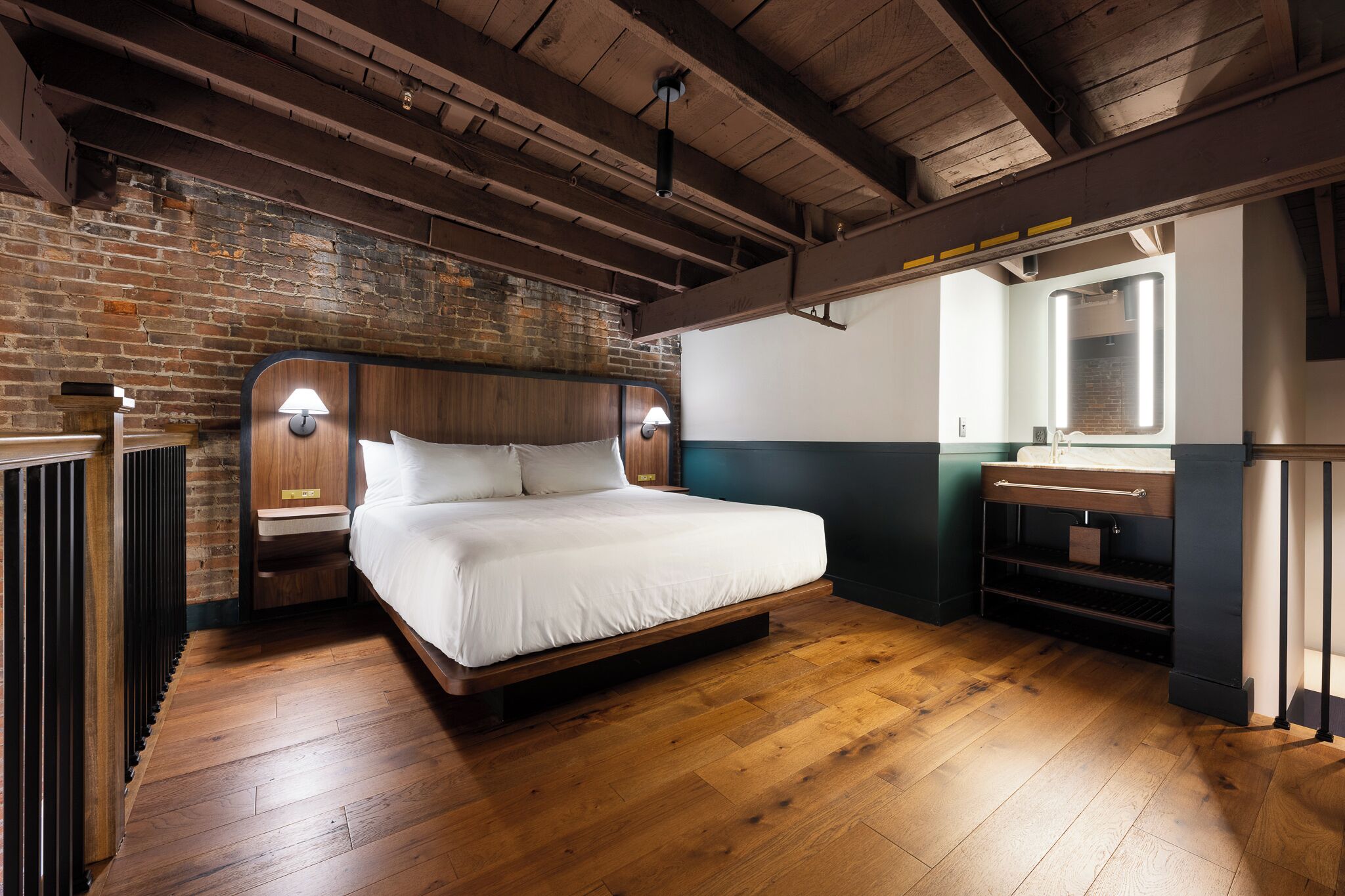 A modern bedroom with a wooden floor, a brick wall, a bed with a wooden headboard, bedside lamps, and a small bedside table.