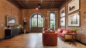 A cozy room with a brick wall, wooden floor, a red sofa, a round ottoman, and a small table with a plant. - The Delegate (Frankfort)