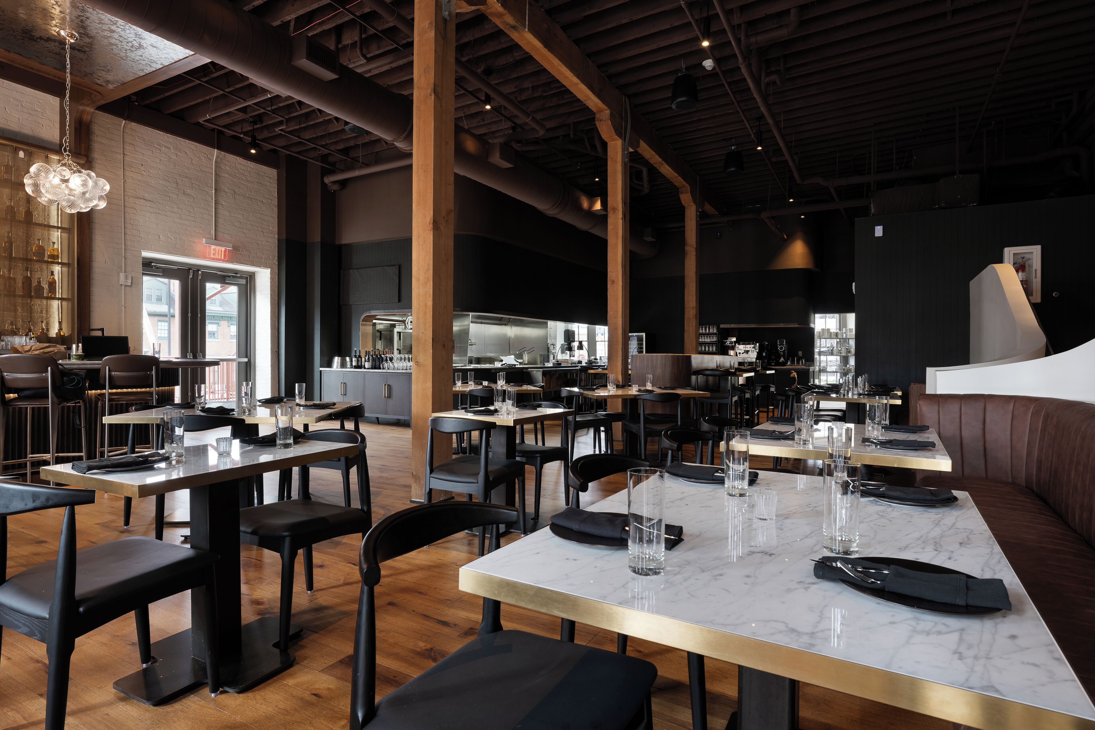 A restaurant with marble-topped tables and black chairs, wooden beams on the ceiling, and a bar area in the background.
