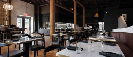 A restaurant with marble-topped tables and black chairs, wooden beams on the ceiling, and a bar area in the background.