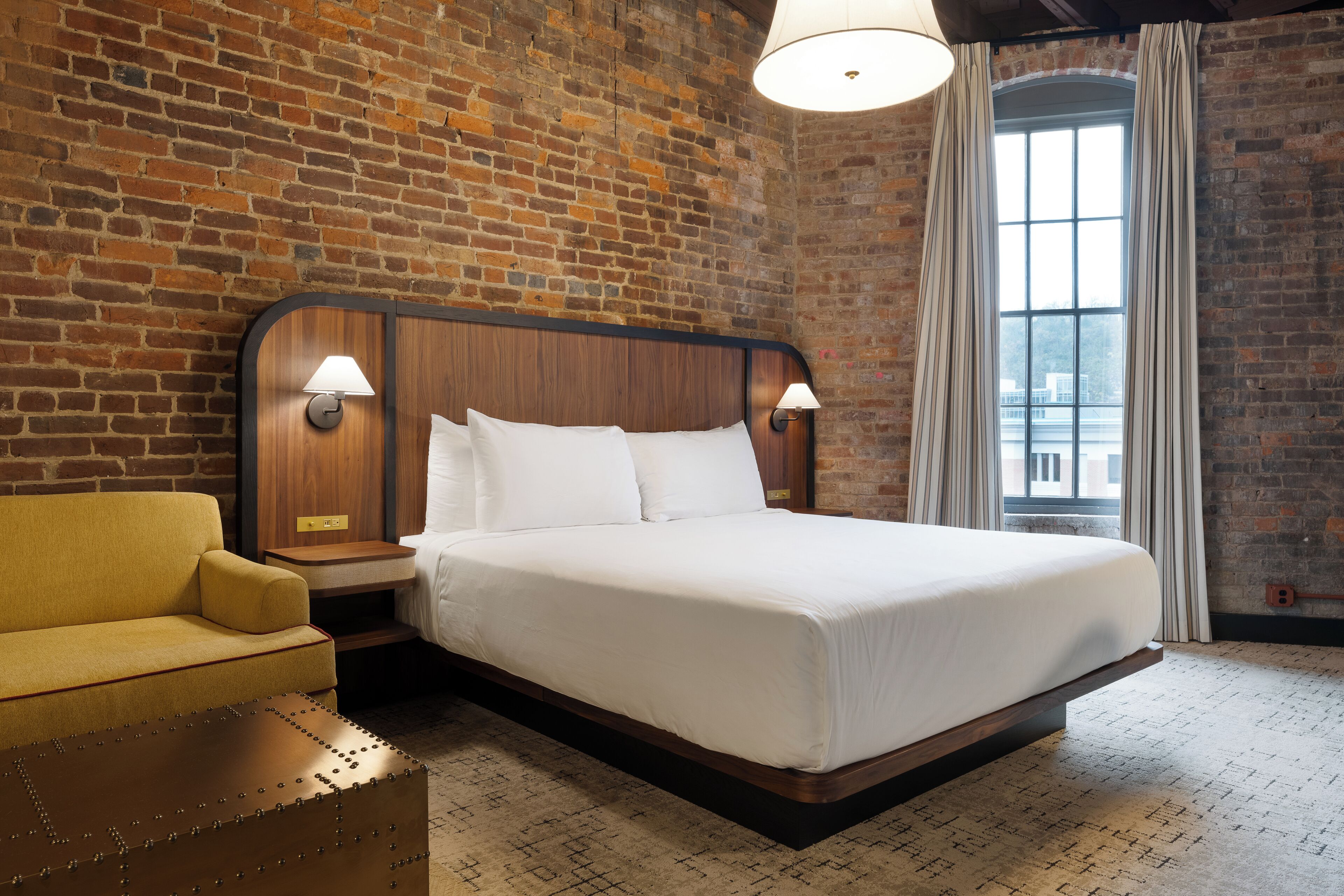 A hotel room with a brick wall, a wooden bed frame, a yellow armchair, and a large window with curtains.