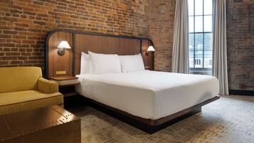 A hotel room with a brick wall, a wooden bed frame, a yellow armchair, and a large window with curtains.