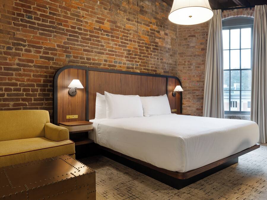 A hotel room with a brick wall, a wooden bed frame, a yellow armchair, and a large window with curtains.