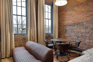 A cozy room with a brick wall, a large window with curtains, a round table with chairs, and a cushioned bench.