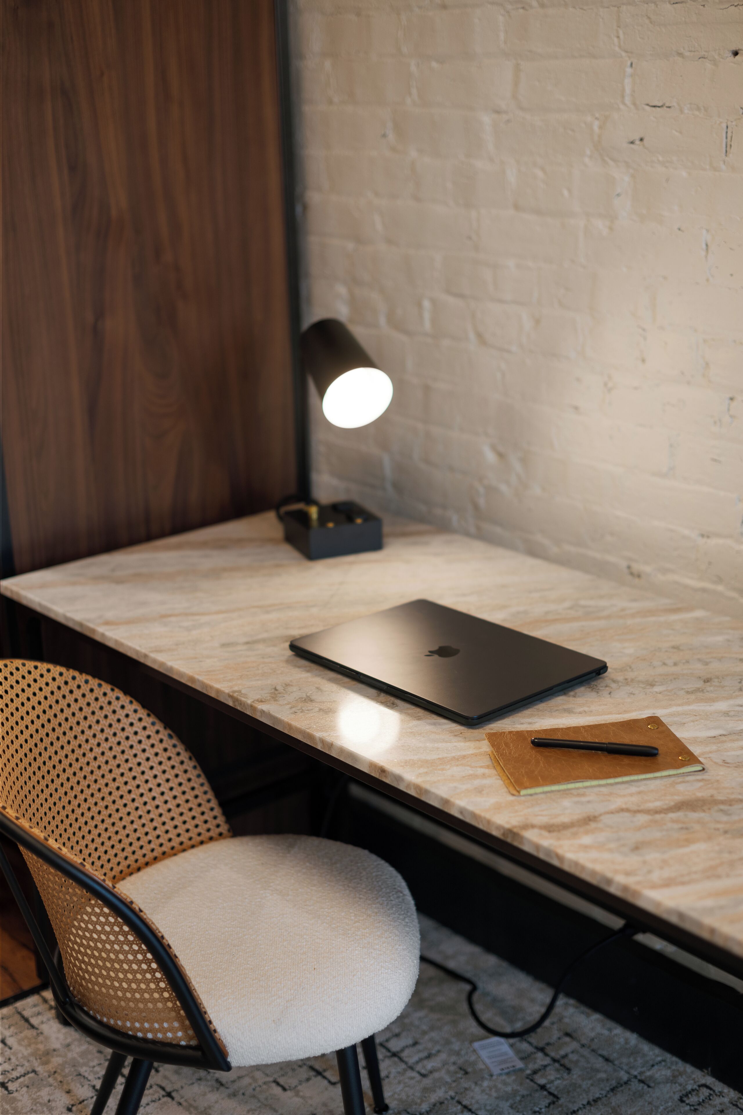 A workspace with a laptop, a notebook, and a pen on a marble table, a chair, and a wall-mounted lamp.