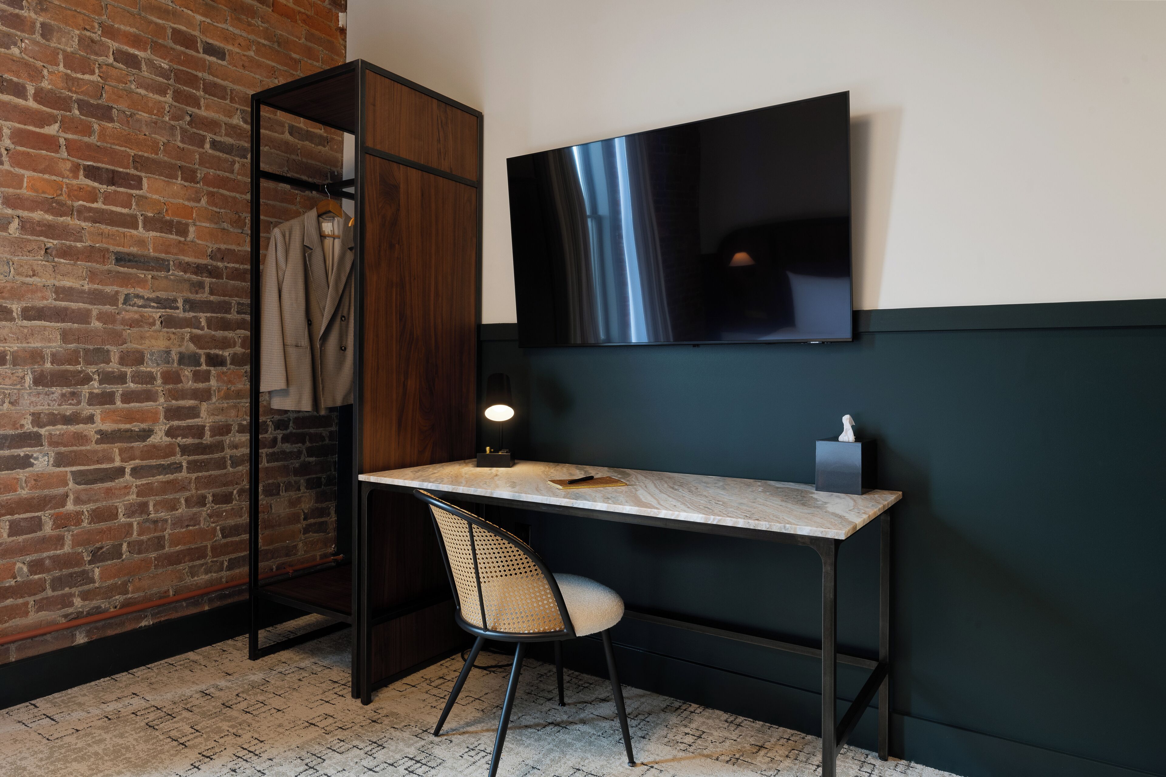 A room with a brick wall, a wooden wardrobe, a desk with a chair, a flat-screen TV, and a small table with a lamp.
