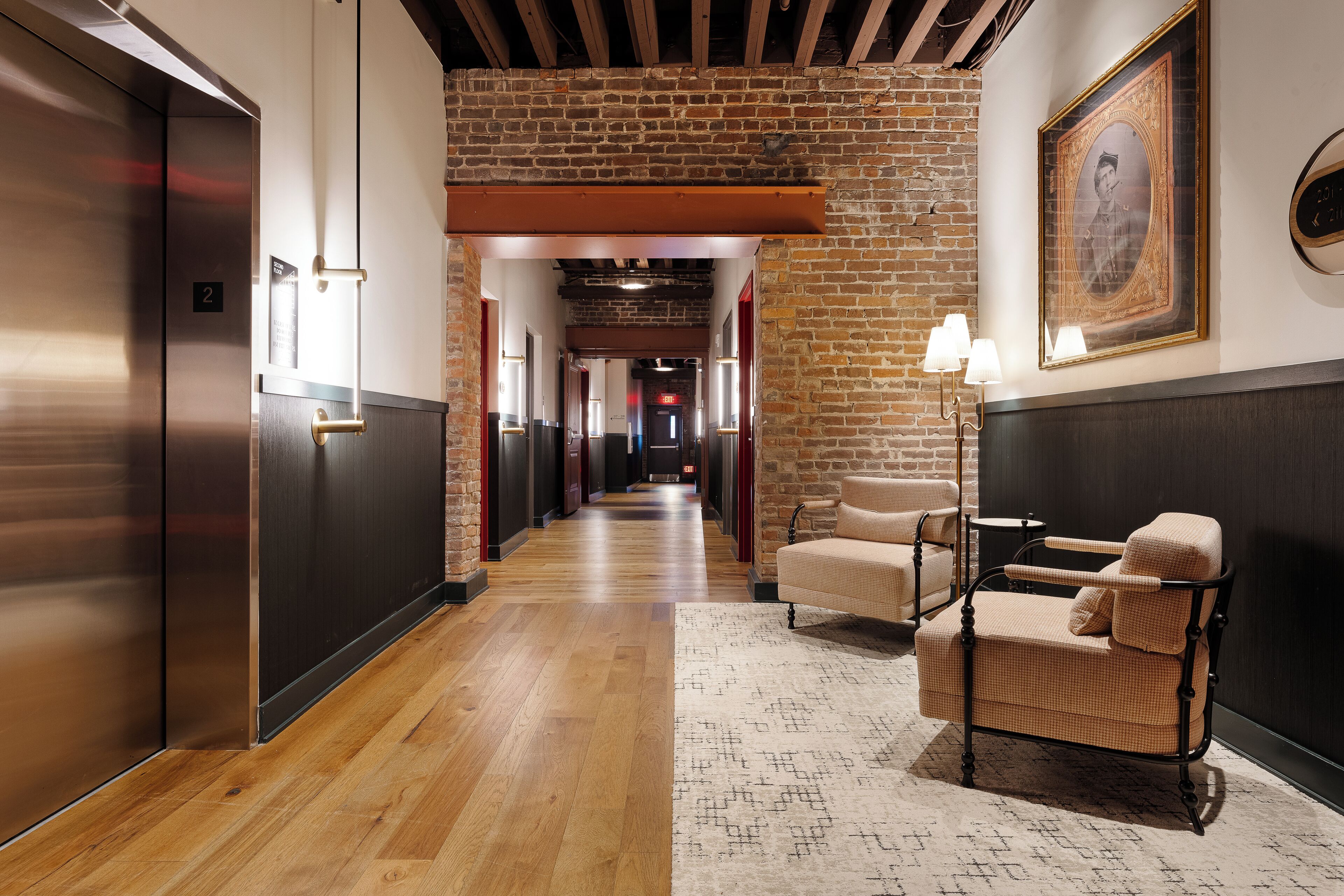 A hotel corridor with a wooden floor, a seating area with a patterned rug, and a framed portrait on the wall.