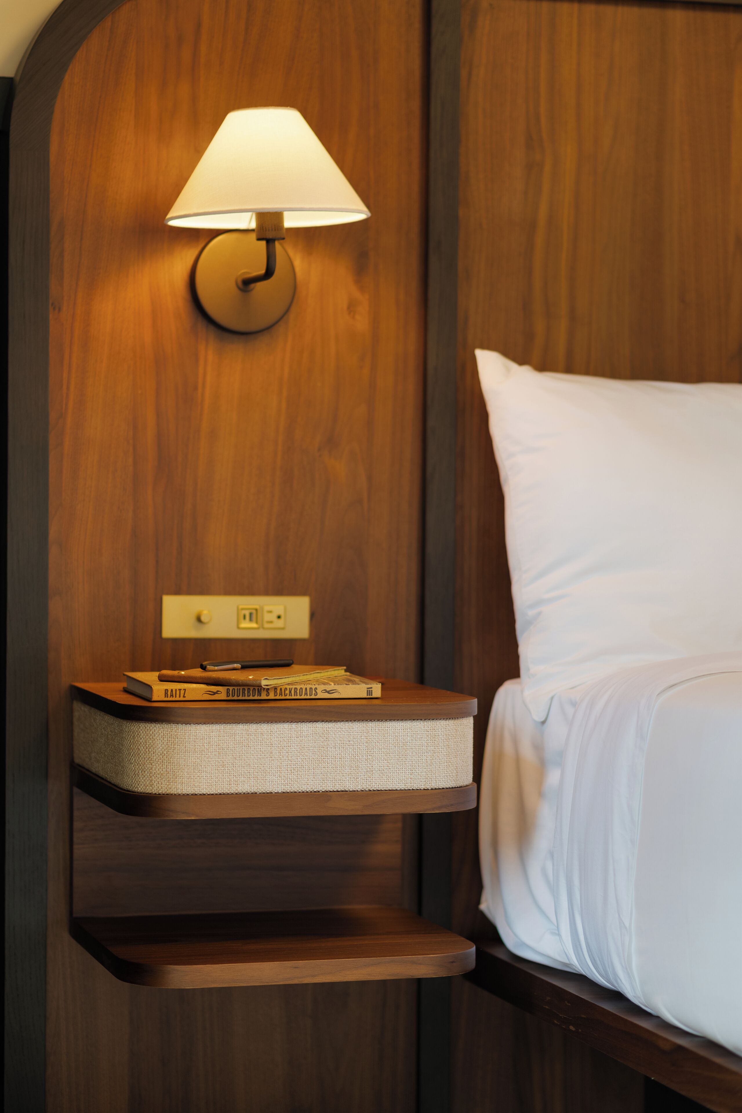 A neatly made bed with white bedding, a bedside lamp, and a small wooden nightstand with books and a pen.