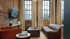 A cozy room with a brick wall, a large window with curtains, a sofa, a coffee table, and a TV mounted on the wall. - The Delegate (Frankfort)