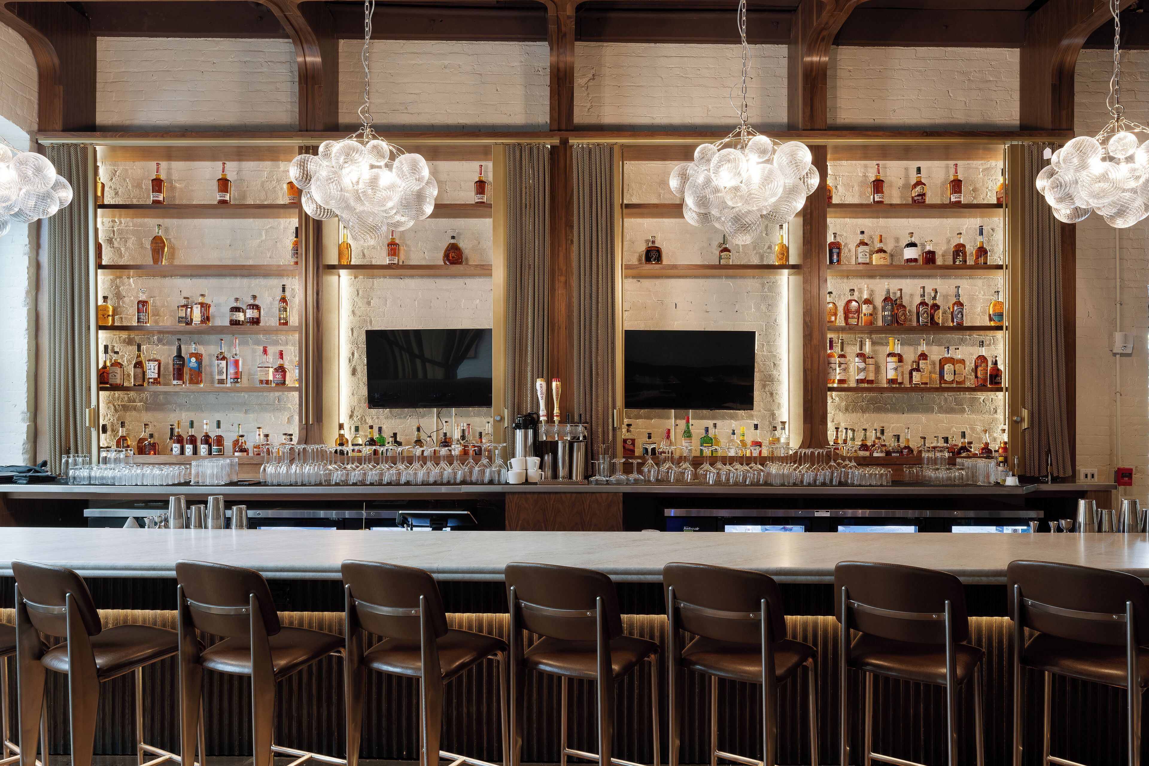 A bar area with multiple shelves of liquor bottles, bar stools, and hanging light fixtures.