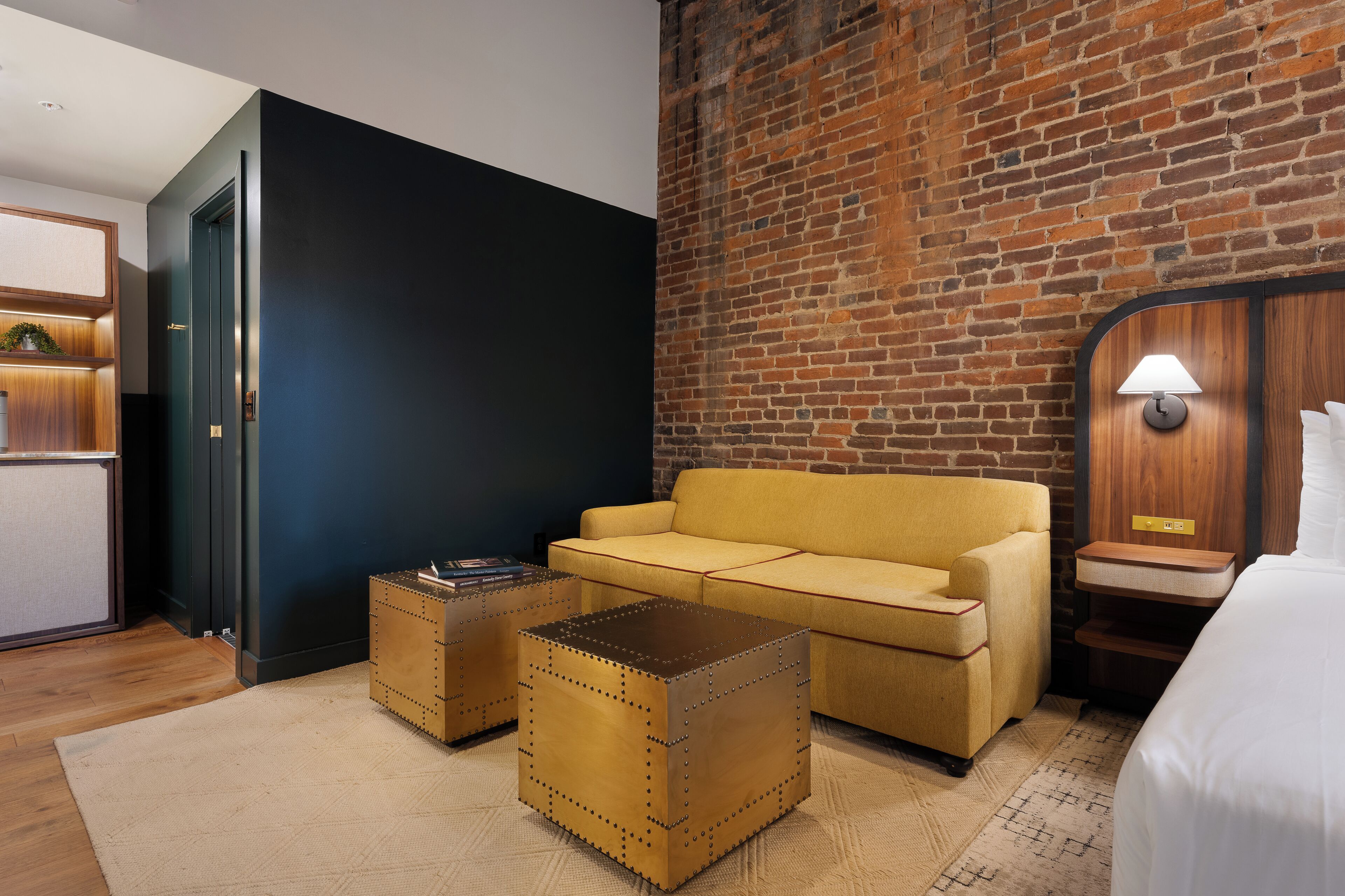 Habitación de hotel con pared de ladrillo, un sofá amarillo, una mesa de centro de madera y una cama con cubrecama blanco.