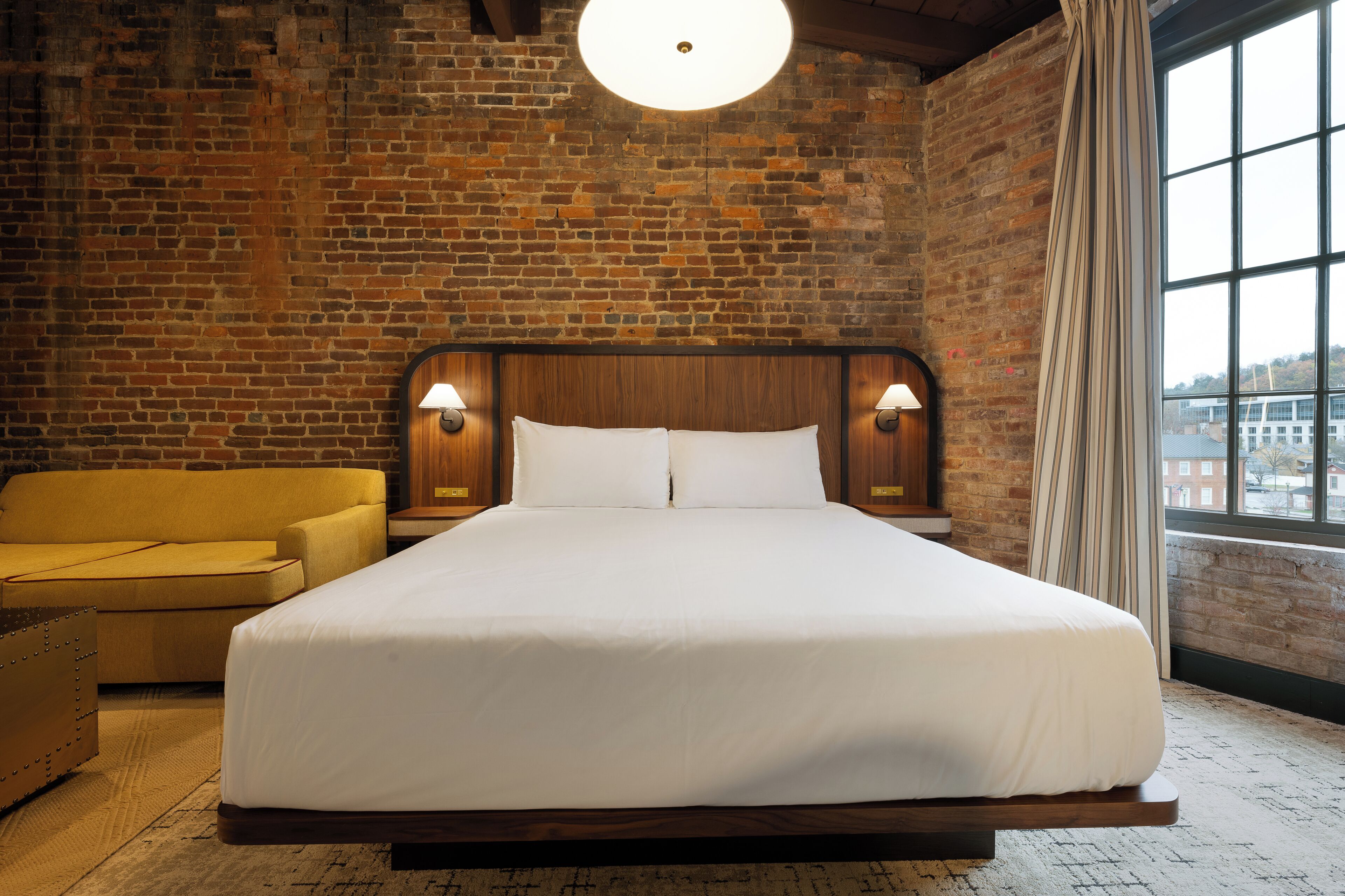 A hotel room with a large bed, a yellow armchair, and a brick wall.