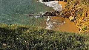 A coastal landscape with a sandy beach, rocky cliffs, and houses in the background.