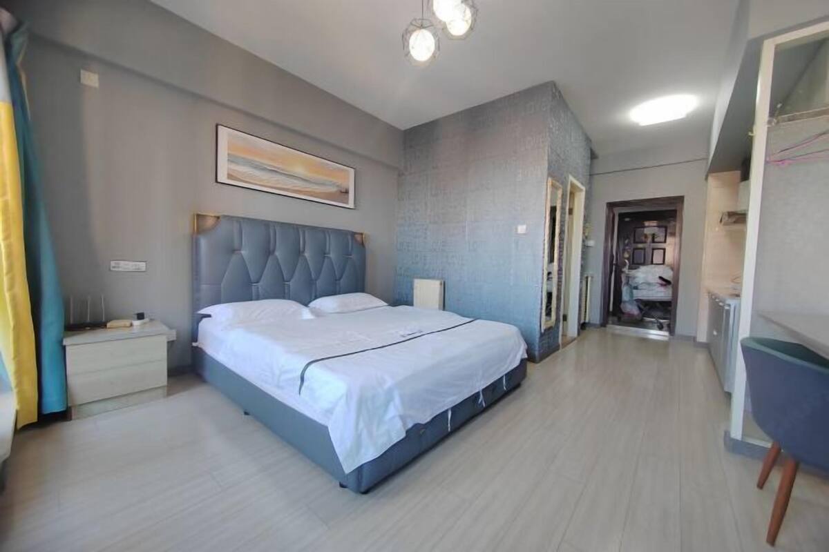 A bedroom with a large bed, a bedside table, a nightstand, a wall-mounted light fixture, and a door leading to another room.
