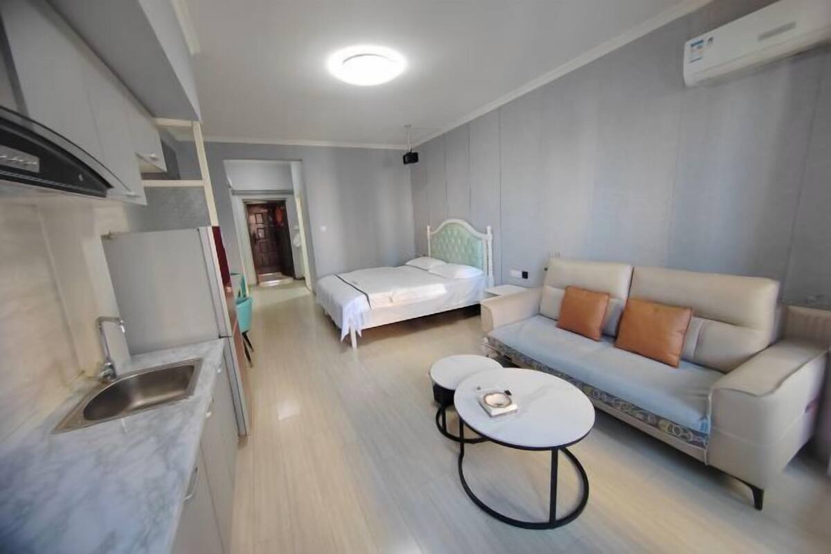 A modern bedroom with a bed, a sofa, a small table, and a kitchen area with a sink and cabinets.