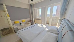 A hotel room with a large bed, a small table, a sofa, and a balcony with a view of the city.