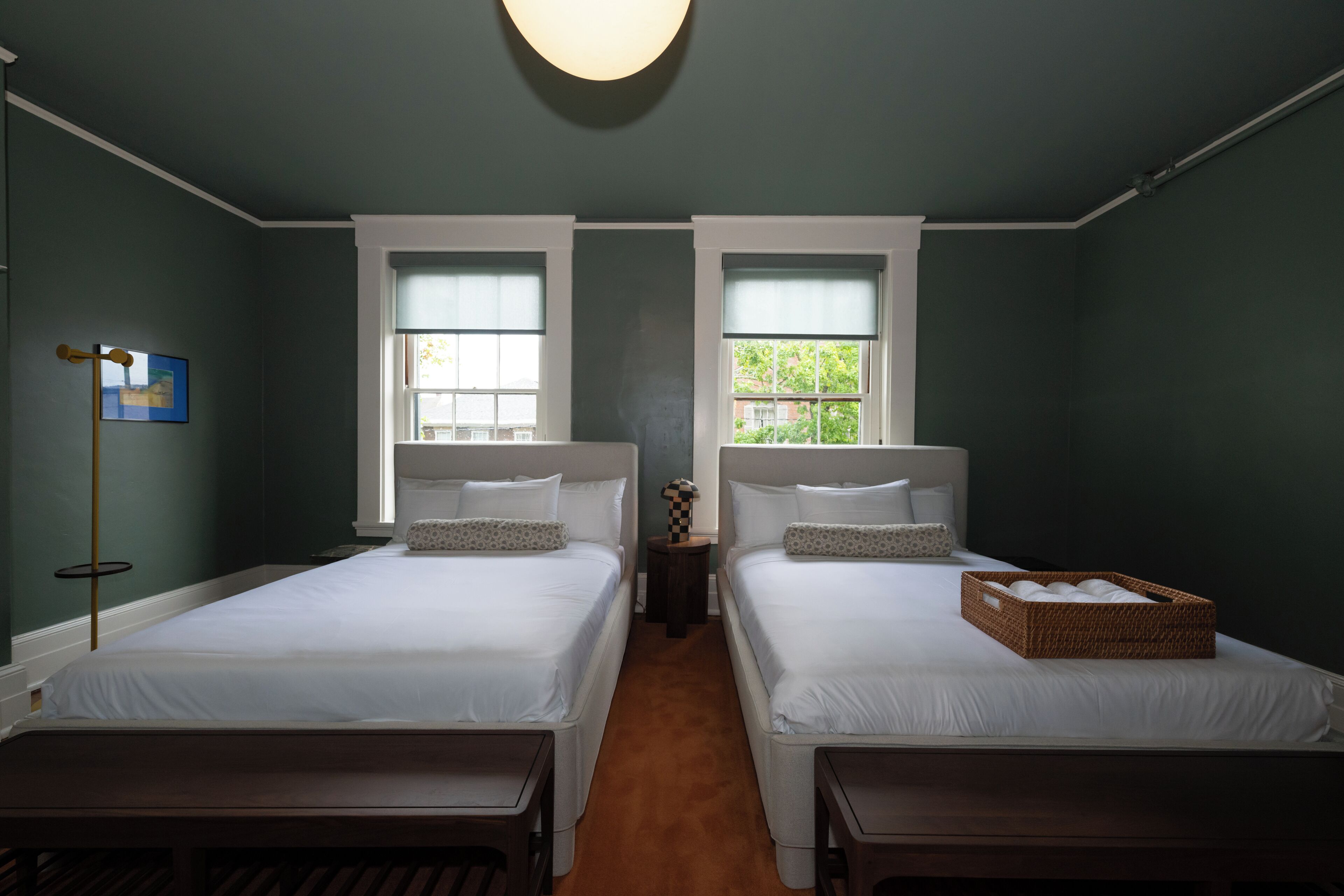 A hotel room with two single beds, a small table, a lamp, and a window with a view of greenery.