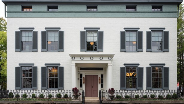 A two-story building with a symmetrical facade, featuring a central entrance with a brown door, flanked by windows with shutters, and a decorative metal fence with potted plants. - The Ashbrook Hotel (Frankfort)