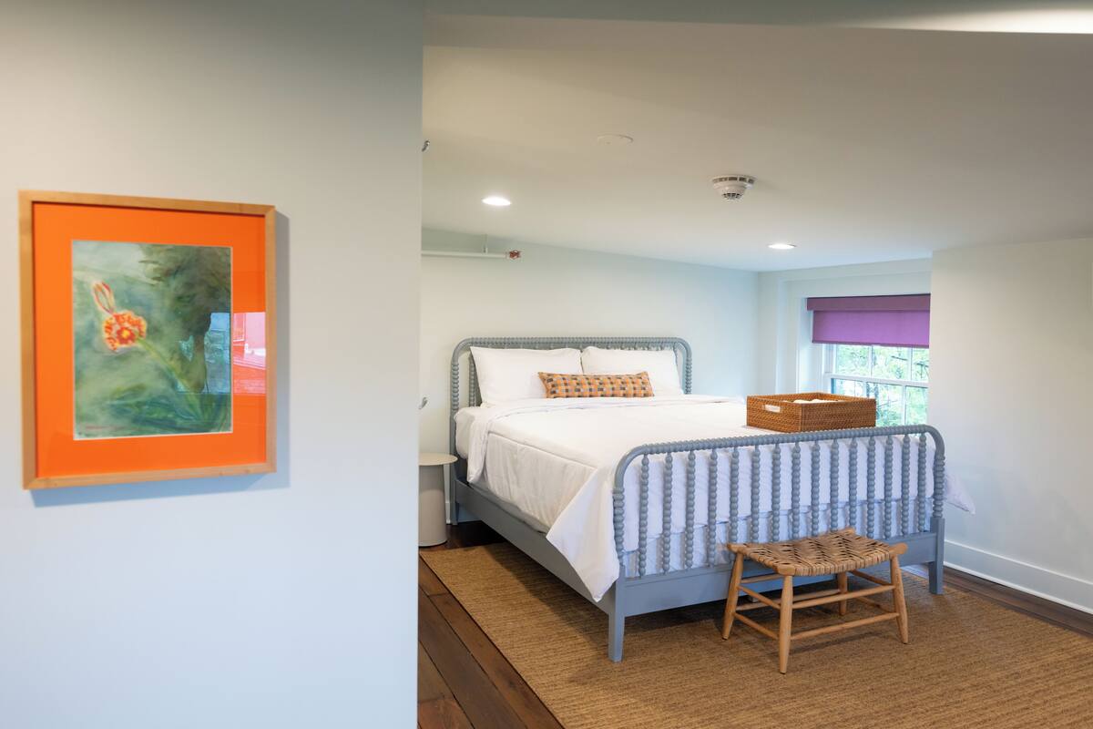A bedroom with a bed, a nightstand, a chair, a window with blinds, and a framed picture on the wall.