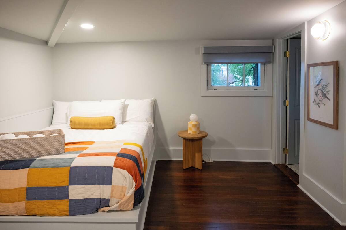 A bedroom with a bed, a window, a small wooden table, and a framed picture on the wall.