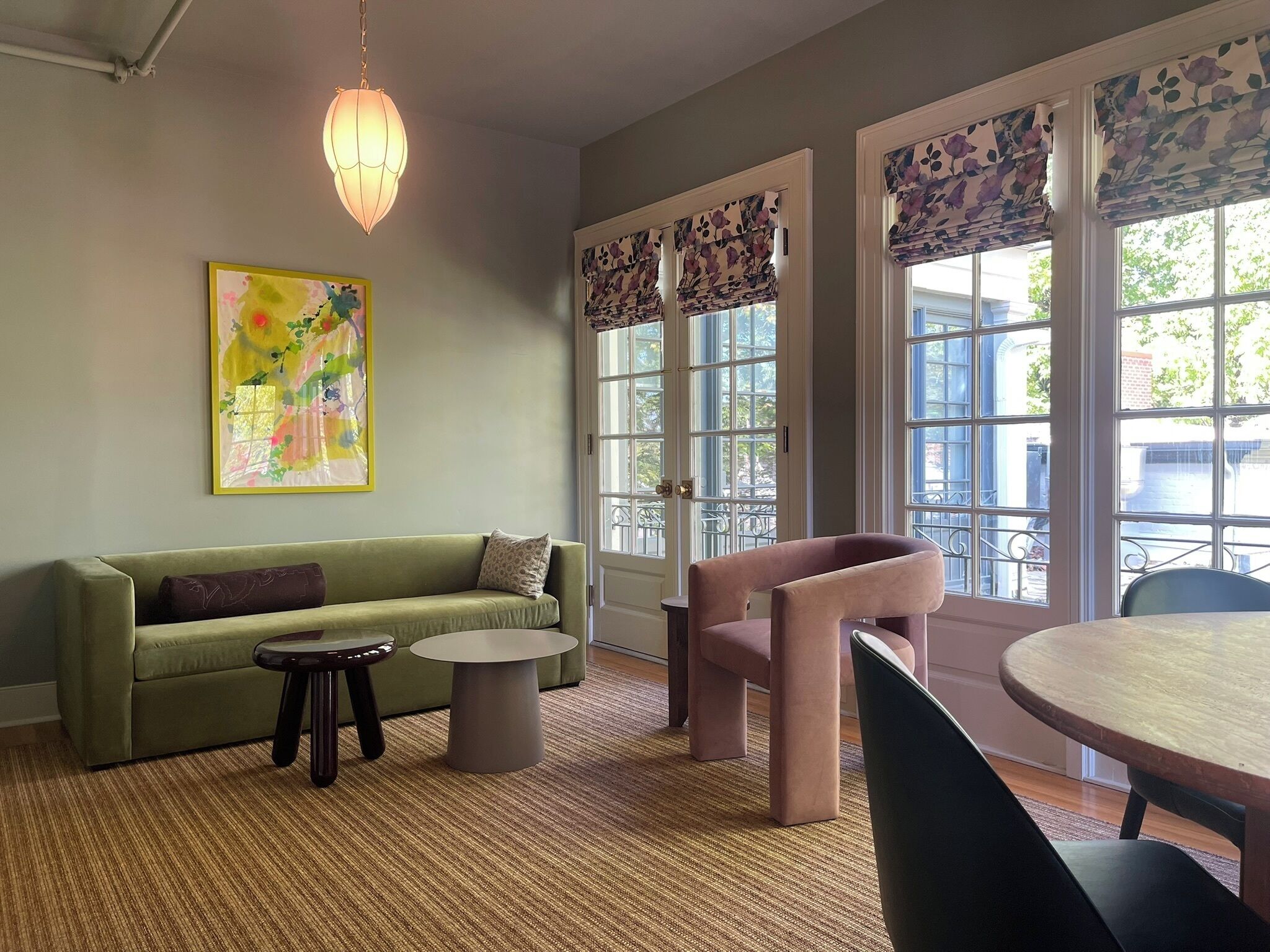 A cozy living room with a green sofa, a round coffee table, and a pink armchair. There is a painting on the wall and a hanging light fixture.