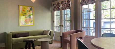 Uma sala de estar aconchegante com um sofá verde, uma mesa de centro redonda e uma poltrona rosa. Há um quadro na parede e um lustre.