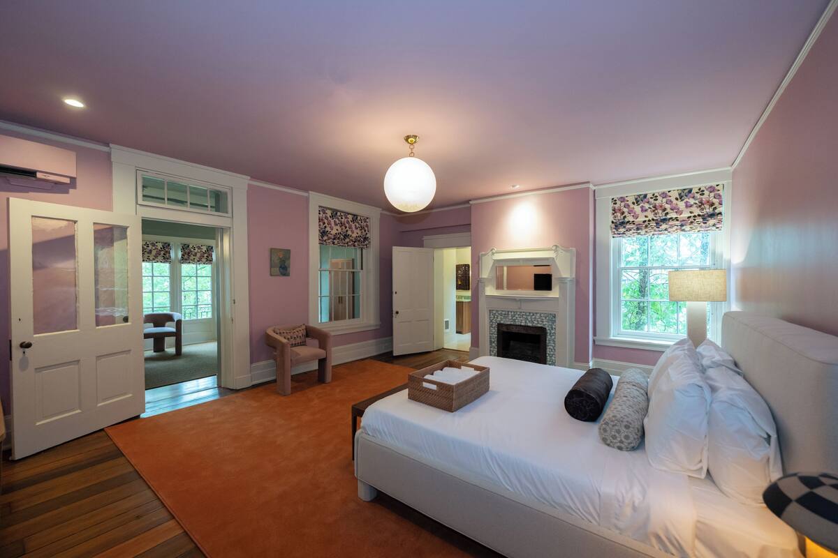 A bedroom with a bed, a fireplace, a door, and a window with floral curtains.