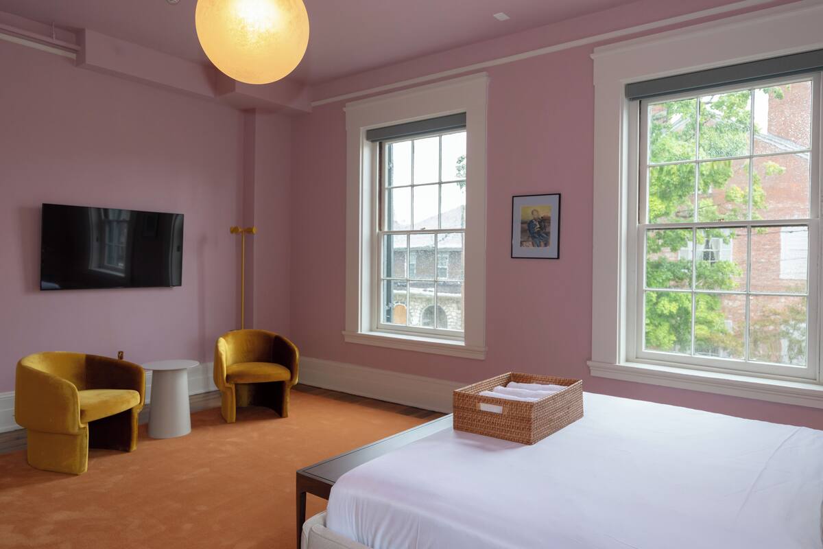 A bedroom with a bed, two chairs, a television, and a window with a view of buildings.