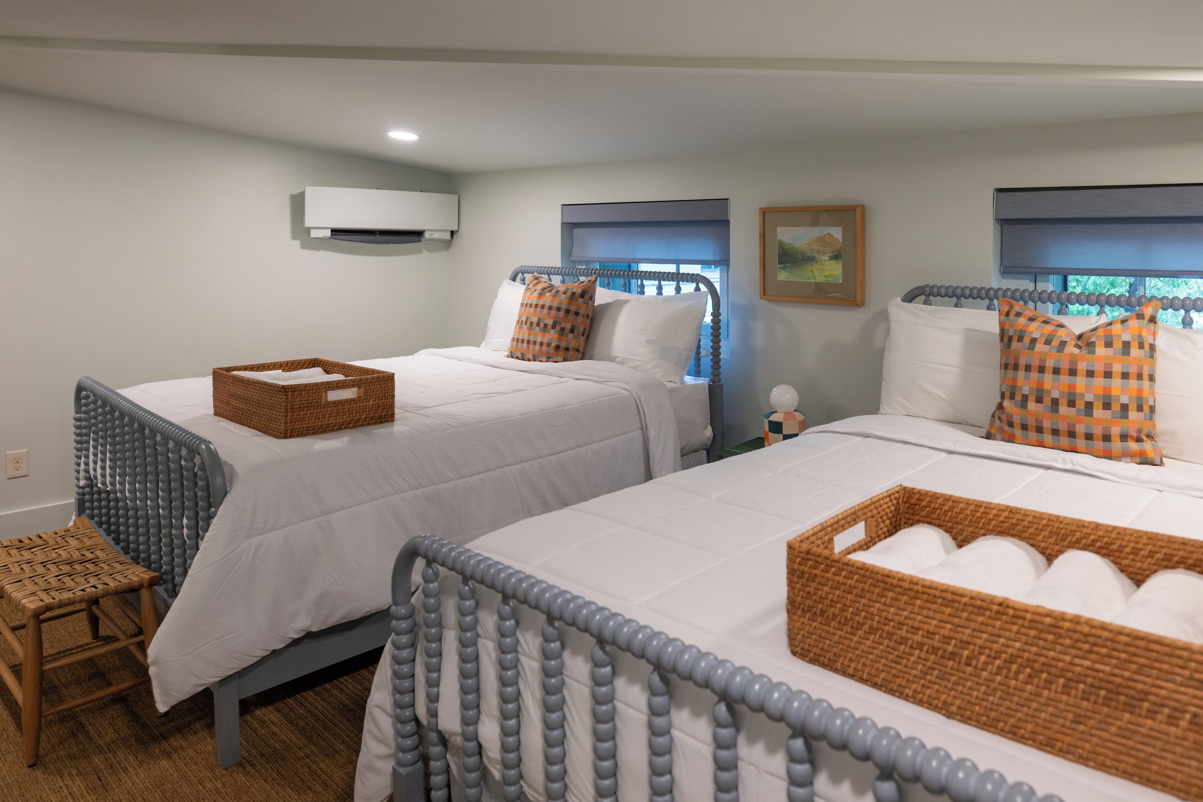 Two twin beds with white bedding and checkered pillows, a wicker basket on each bed, a small wooden stool, and a wall-mounted air conditioner.