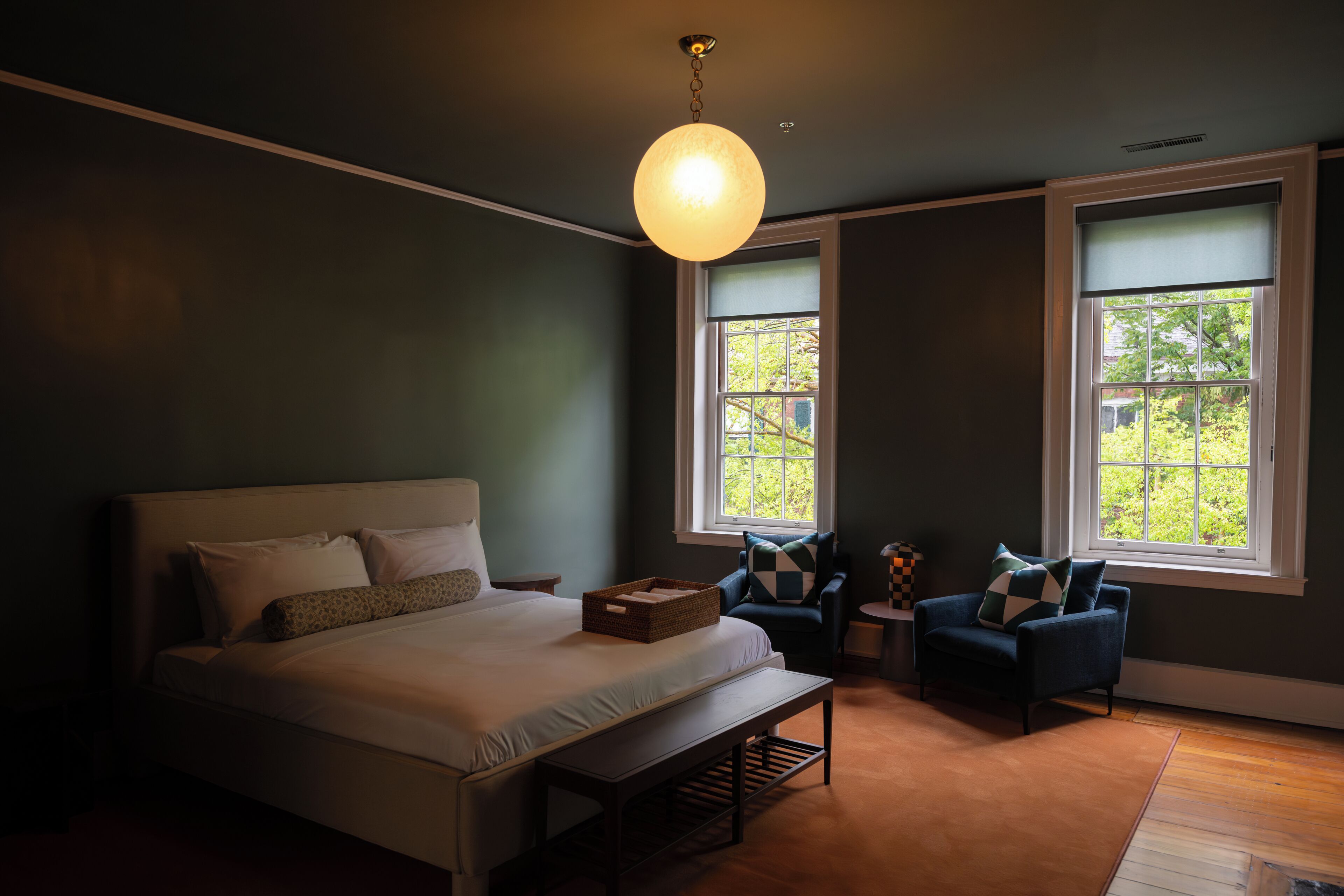 A bedroom with a large bed, two armchairs, a small table, and two windows with views of greenery.