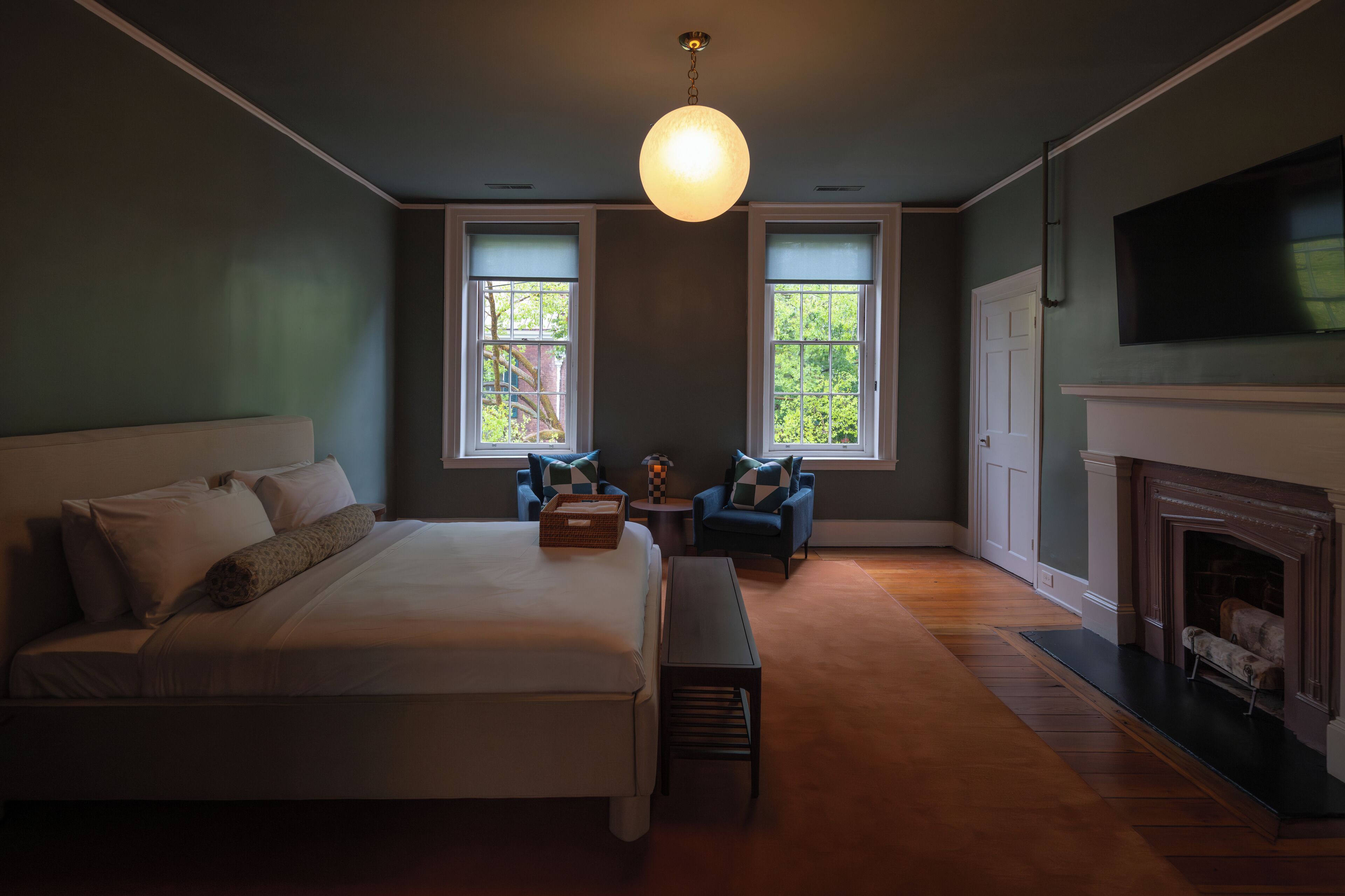 A bedroom with a large bed, two armchairs, a fireplace, and two windows with views of greenery.