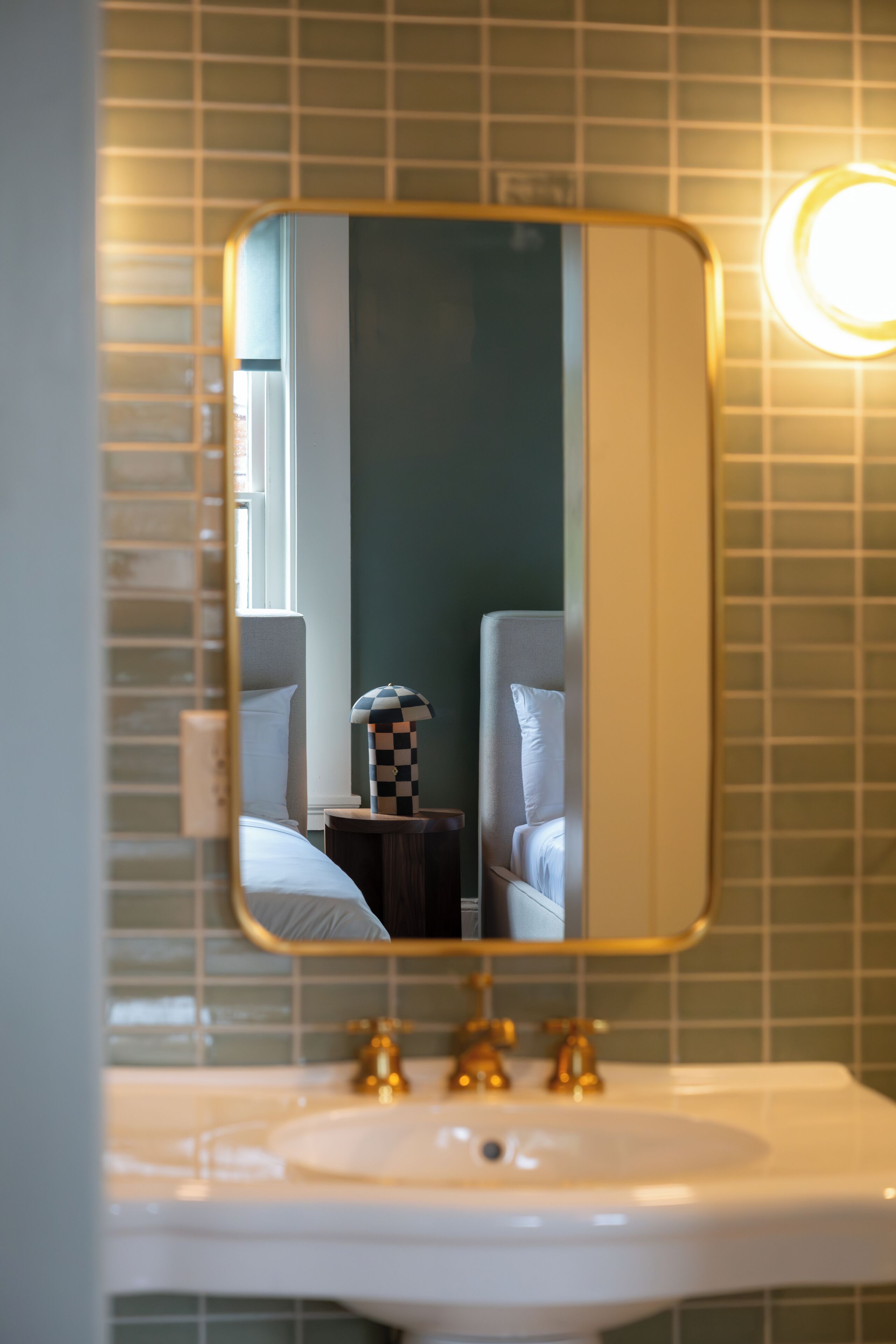 Une salle de bain avec un lavabo blanc, des robinets dorés et un miroir qui reflète un lit surmonté d’une lampe à motifs à carreaux.