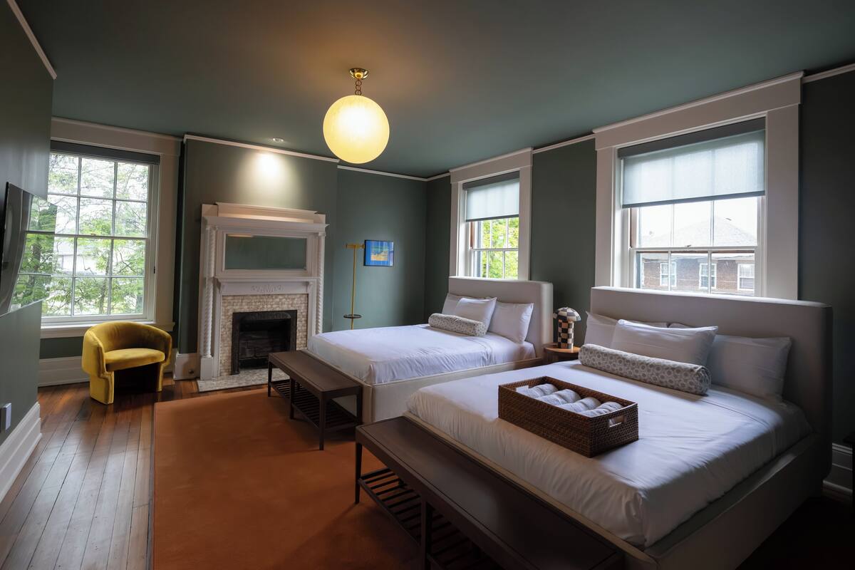 A bedroom with two beds, a fireplace, a yellow armchair, and large windows.
