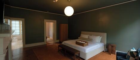 A bedroom with a large bed, a bedside table, a bench, a small wooden stool, and a view of a hallway.