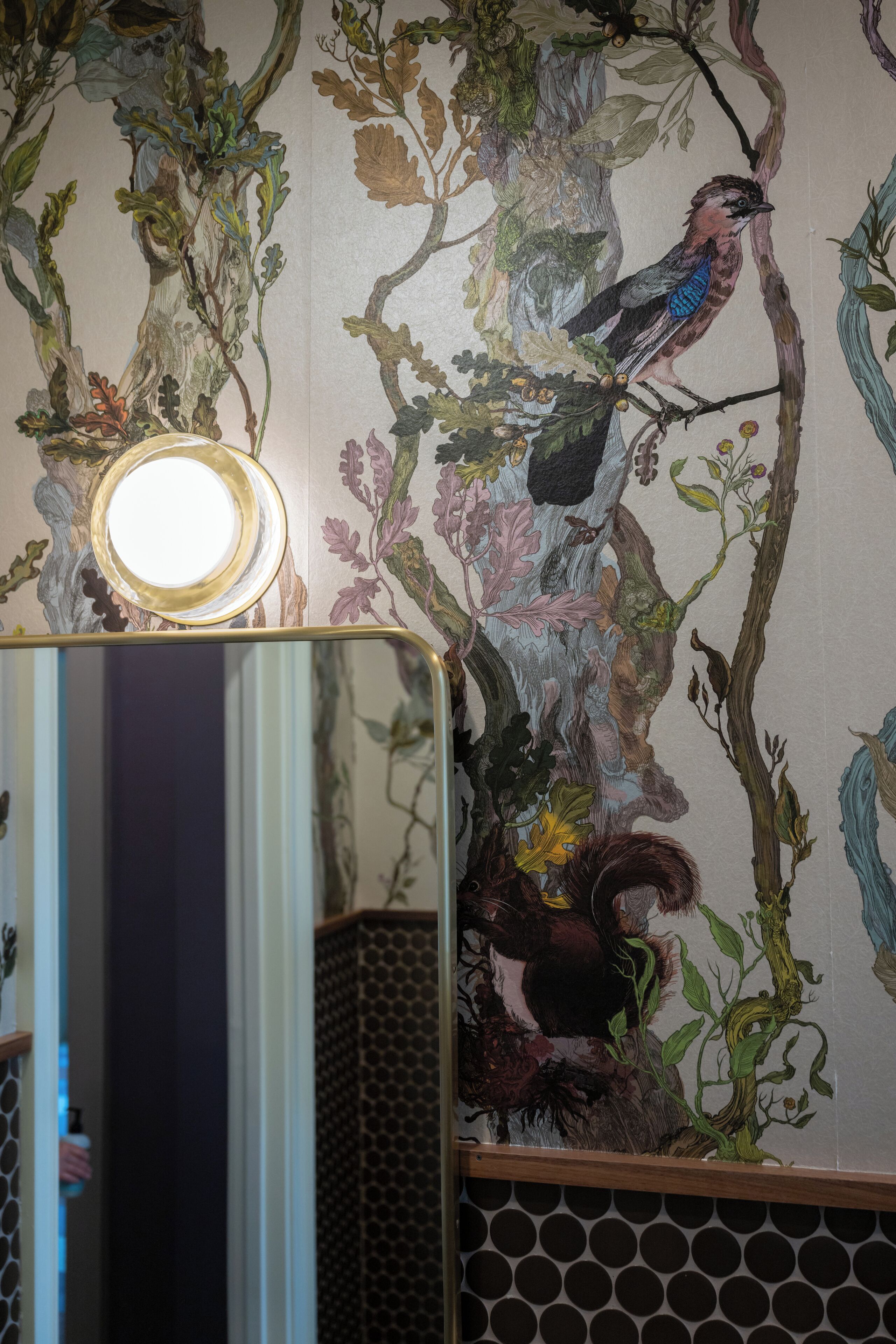 A room with a wall mural of a bird perched on a branch, a round mirror with a light above it, and a patterned tile floor.