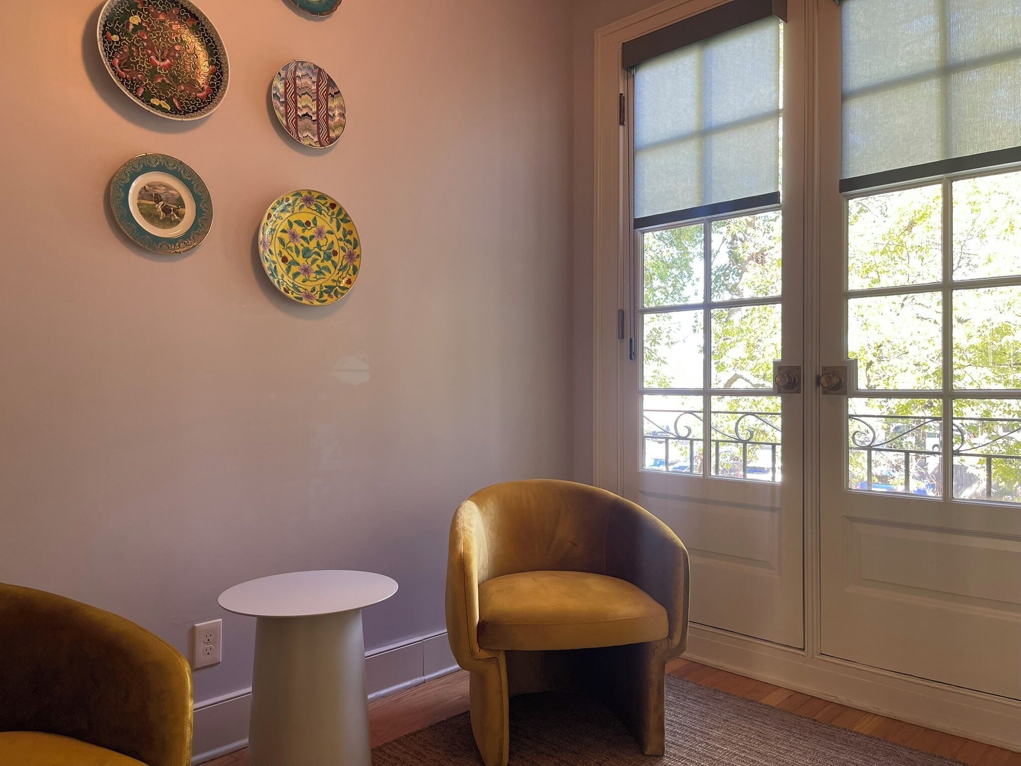 A room with a large window, a door, and a wall decorated with four plates. There is a small round table and a chair in the room.